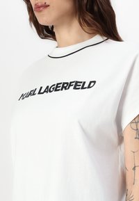 Kreis balts kokvilnas t-krekls ar melnu uzrakstu "KARL LAGERFELD", īsām piedurknēm un melnu apmalojumu apkaklē. Gluda tekstūra ar brīvu piegriezumu.