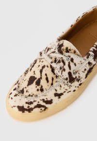 Gros plan sur une chaussure mocassin tannée à taches marron foncé avec une semelle en caoutchouc brun clair et des lacets assortis sur un fond blanc.