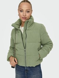 Veste en velours côtelé vert avec un col montant et une fermeture éclair à l'avant, présentant une surface texturée et des poches latérales, portée sur un haut blanc.