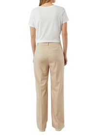 Kobieta zwrócona tyłem, ubrana w białą koszulkę z krótkim rękawem włożoną do beżowych, wysokiej talii, szerokich spodni z tylną kieszenią.