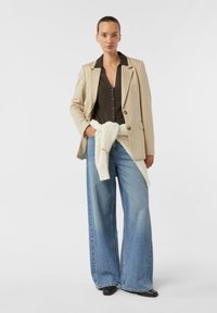 Gestreifter beiger Blazer über einem dunkelknöpfbaren Hemd, kombiniert mit hellblauen, weit geschnittenen Jeans und einem weißen Strickpullover, der in der Taille gebunden ist. Schwarze Schuhe.