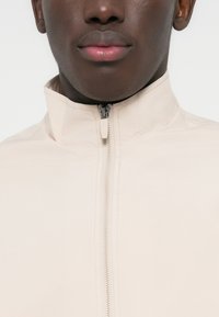 Κοντινό πλάνο του κάτω μέρους του προσώπου και του λαιμού ενός ατόμου που φοράει μπεζ μπουφάν με φερμουάρ στον ψηλό γιακά, μπροστά σε λευκό φόντο.