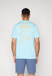 Φωτεινό γαλάζιο βαμβακερό t-shirt με πορτοκαλί κείμενο "ADIDAS PADEL" και γραφικό γήπεδου padel στην πλάτη. Κοντά μανίκια, στρογγυλή λαιμόκοψη.