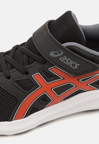 Black athletic shoe with mesh upper, red and white accents, velcro strap, and padded collar. Features textured sole for traction.