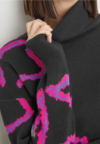 Svart tröja med hög hals och långa ärmar, prydd med ett texturerat abstrakt mönster i rosa och lila. Mjuk stickad tyg.