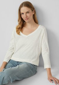 Crème-kleurige lange mouwen V-hals shirt van een zachte, lichtgewicht stof, gecombineerd met lichtblauwe jeans, zittend tegen een neutrale achtergrond.