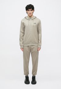 Lys beige hettejakke med glidelås og snører, med logo, kombinert med matchende avsmalnende joggebukser, brukt med svarte sportssko. Jevn tekstur.