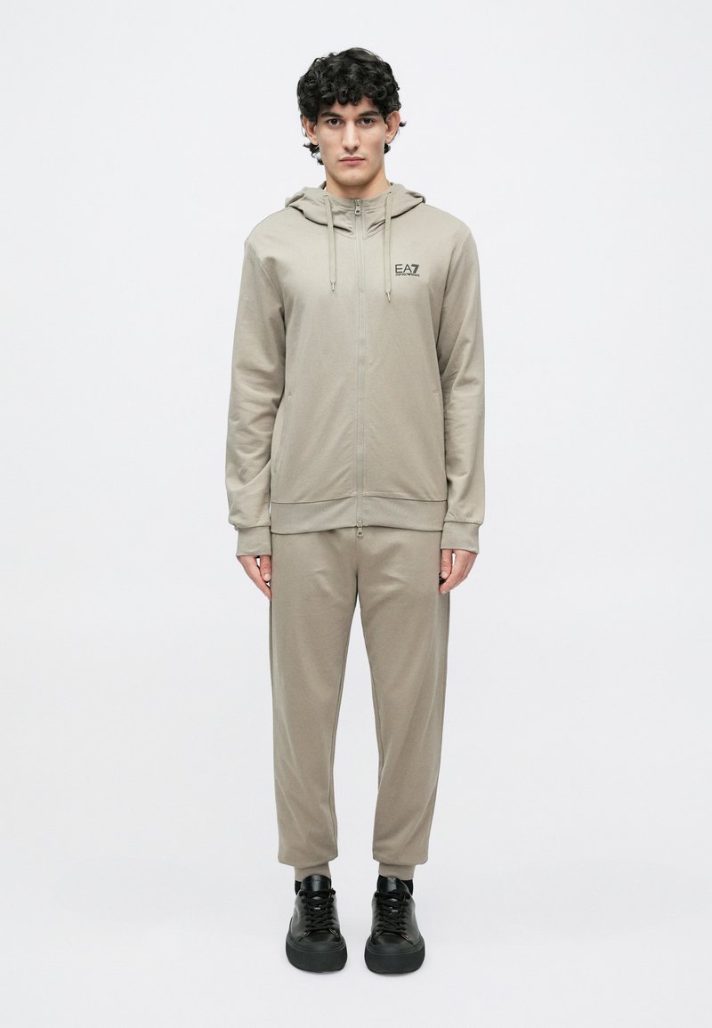 Lys beige hettejakke med glidelås og snører, med logo, kombinert med matchende avsmalnende joggebukser, brukt med svarte sportssko. Jevn tekstur.