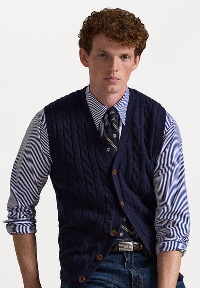 Marineblå kabelstrikket vest med brune knapper, båret over en hvit og blåstripet skjorte. Har et slips med blå og gullmønster.