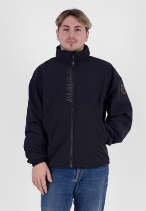 Giacca nera con zip e colletto alto, tessuto strutturato e patch con logo sulla manica, abbinata a jeans blu.