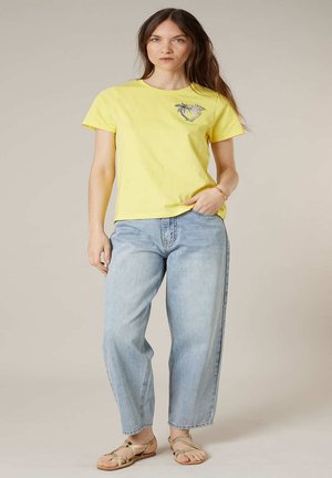 Femme portant un t-shirt jaune avec un dessin de palmier, un jean bleu clair et des sandales beige, debout devant un fond uni.
