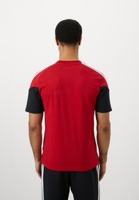 Homem de costas a usar uma camisa de manga curta vermelha e preta com riscas brancas nos ombros e calças pretas com riscas brancas laterais.