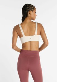 Sujetador deportivo beige claro con tirantes ajustables, con un logo sutil en la parte trasera. Combinado con leggings de talle alto en color granate.