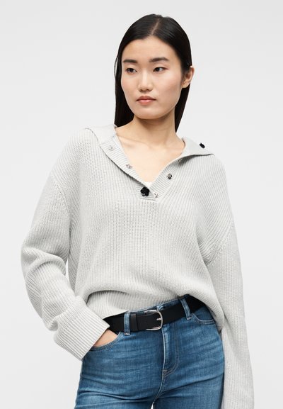 Ljusgrå stickad tröja med V-ringning, stor krage och svarta knappdetaljer, kombinerad med blå denimjeans och ett svart bälte.