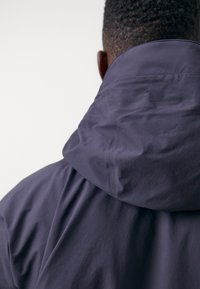 Dark purple hooded jacket with visible stitching, a soft texture, and gathered fabric at the back, showcasing a minimalist design.