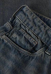 Povečava temno modrih jeans kavbojk, ki prikazuje sprednji žep, kovancev žep, zanko za pas in kovinski zatič.