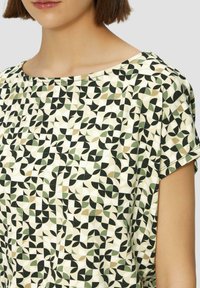 Korte mouwen top in lichtgewicht stof, met een geometrisch patroon in groen, zwart en beige op een crèmekleurige achtergrond. Ronde halslijn.
