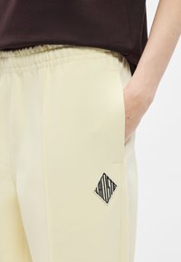 Ανοιχτό κίτρινο παντελόνι κατασκευασμένο από υφή υλικού· διαθέτει ελαστική μέση και μαύρο λογότυπο σε σχήμα διαμαντιού στην τσέπη.