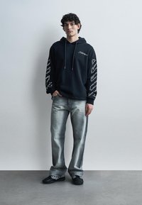 Νέος άνδρας στέκεται σε έναν λευκό τοίχο, φοράει μαύρο φούτερ με λευκό κείμενο στα μανίκια και ανοιχτό γκρι τζιν με μαύρα αθλητικά παπούτσια.