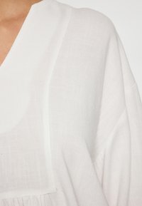 Bílý lněný oděv s volným střihem, s výstřihem do V, jemnou texturou a nenápadným tkaným vzorem. Důraz je kladen na detail lemování.
