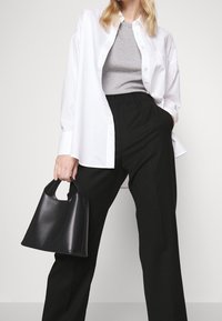 Chemise blanche à boutons portée sur un top gris, associée à un pantalon noir taille haute. Tenant un sac à main en cuir noir structuré.