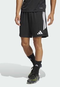 Κάτω σώμα ενός ατόμου που φορά μαύρες αθλητικές βερμούδες Adidas, μαύρες κάλτσες με λευκά λογότυπα και μαύρα και πράσινα παπούτσια ποδοσφαίρου σε ανοιχτό φόντο.