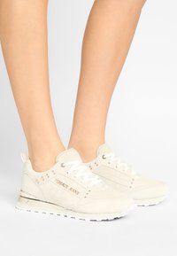 Baskets en daim beige clair avec semelle blanche, lacets à l'avant, accents embellis et logo sur le côté.