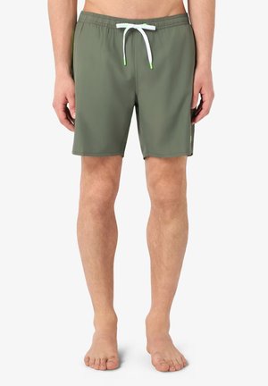 Uomo con pantaloncini da bagno verde oliva con coulisse bianca, in piedi a piedi nudi contro uno sfondo bianco semplice.