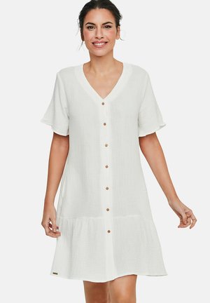 Femme souriante portant une robe chemise blanche à manches courtes avec un col en V et un ourlet volanté, debout devant un fond uni.