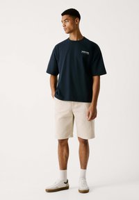 Dunkelblaues Baumwoll-T-Shirt mit kurzen Ärmeln und lockerer Passform, kombiniert mit hellbeigen Baumwollshorts und weißen Sneakers mit braunen Sohlen.