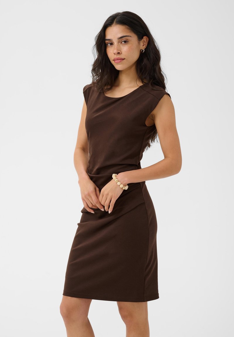 Femme aux longs cheveux foncés portant une robe marron sans manches jusqu'aux genoux et un bracelet beige à perles, debout devant un fond uni.