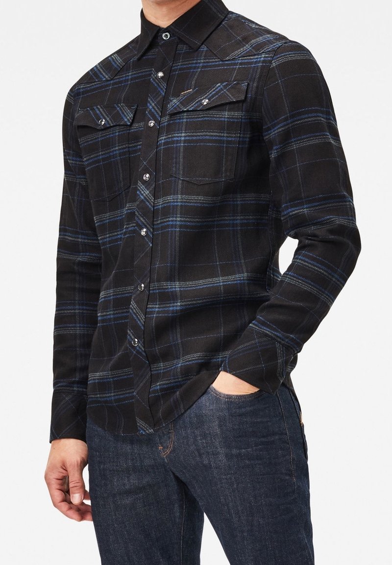 Camisa de cuadros negro y azul de manga larga con collar abotonado, dos bolsillos en el pecho y detalles de botones plateados, llevada sobre jeans de mezclilla oscuros.