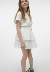 Vestido blanco de capas con acabado texturizado y mangas cortas. Presenta detalles de botones y ribete de encaje. Se lleva con zapatillas verdes y una pulsera de perlas.