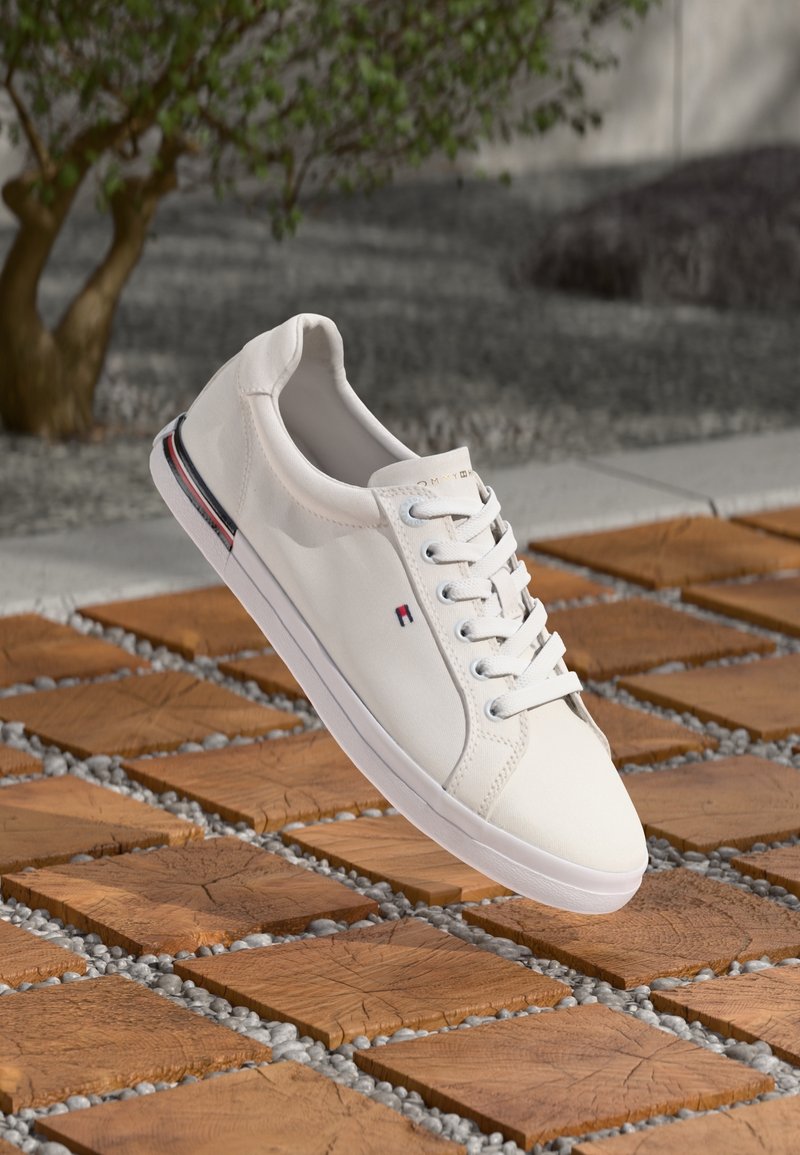 Baskets en toile blanche avec lacets plats, profil bas et semelle en caoutchouc. Elles présentent une bande d'accent rouge et bleue ainsi qu'un logo sur le côté.