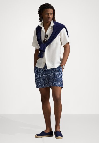 Lys kortermet skjorte i lys beige, blomstrete navy shorts, navy genser over skuldrene, og blå espadrillos.