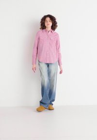 Chemise à carreaux rouge et blanche, jean clair délavé, chaussures beige, se tenant sur un fond blanc. Deux poches poitrine visibles.