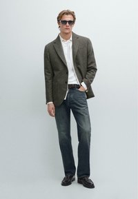Blazer grigio testurizzato sopra una camicia bianca, abbinato a jeans blu scuro e mocassini neri. Occhiali da sole completano il look.