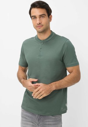 Uomo dai capelli scuri che indossa una polo verde a maniche corte e jeans grigi chiari, in piedi con le mani incrociate all'altezza della vita.