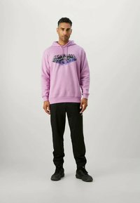 Ανοικτό μωβ φούτερ με εκτύπωση γραφικών, συνδυασμένο με μαύρα παντελόνια φόρμας και μαύρα αθλητικά παπούτσια. Το ύφασμα φαίνεται απαλό και υφασμένο.