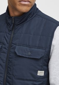 Marineblaue gesteppte Weste mit hohem Kragen, Reißverschluss und knöpfbaren Brusttaschen. Mit Markenlabel an der Seitennaht und strukturiertem Stoff.