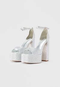 Srebrne petke z visokim podplatom, okrašene z dragocenimi kamni na zgornjem delu, odprtimi prsti in paščkom okoli gležnja, s kockasto peto in kontrastnimi teksturiranimi poudarki.