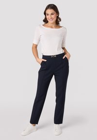 Marineblauwe op maat gemaakte broek met zijzakken en een gouden accent-belt, gecombineerd met een witte bootneck top en witte sneakers. Gladde stof.