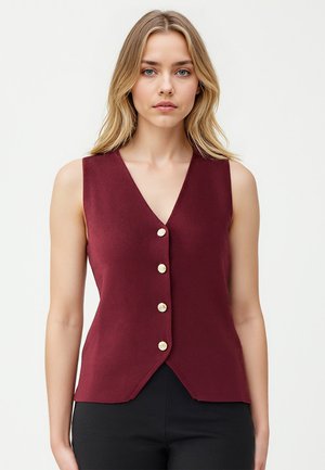 Gilet sans manches bordeaux avec un col en V, comportant cinq boutons ronds dorés sur le devant et un ourlet légèrement asymétrique. Tissu texturé.