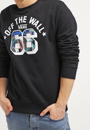 Homme portant un sweat-shirt Vans noir avec l'inscription « Off The Wall 66 » et un motif tropical, les mains dans les poches, associé à un jean bleu.