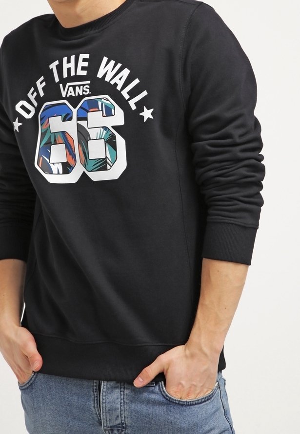 Homme portant un sweat-shirt Vans noir avec l'inscription « Off The Wall 66 » et un motif tropical, les mains dans les poches, associé à un jean bleu.