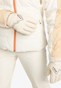 Krémové rukavice s brandingom Roxy, s textúrovaným povrchom a cik-cak detailmi, nosené s bielou a broskyňovou bundou.