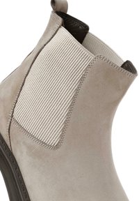 Botín de tobillo de ante beige con paneles laterales elásticos acanalados, tobillo acolchado y suela de goma oscura. Presenta detalles de costura y una pestaña para tirar.