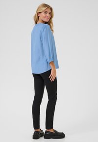 Lichtblauw, lange mouwen top in een zachte stof met pofmouwen. Gecombineerd met zwarte slim-fit broeken en zwarte schoenen. Achteraanzicht getoond.