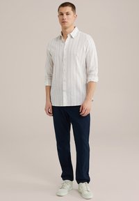 Bela lanena srajca s gumbi in finimi navpičnimi črtami, v kombinaciji s temno modrimi hlačami in belimi supergami. Ohlapen kroj, dolgi rokavi.