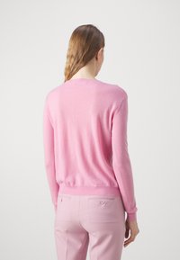 Różowy sweter robiony na drutach z okrągłym dekoltem, długimi rękawami i ściągaczem. Ustawiony na jednolitym tle, tworzy miękki, teksturowany wygląd.